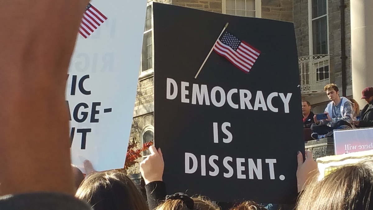 black sign with a flag at the top. Underneath the flag are the words, "Democracy is Dissent."