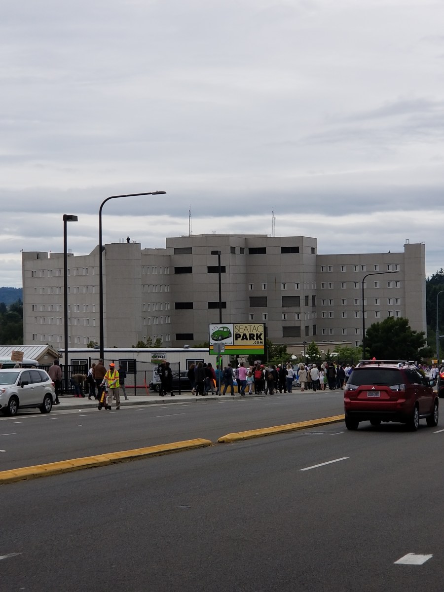 picture of the Federal Detention Center with cars driving down street and people walking towards the rally in front of the building